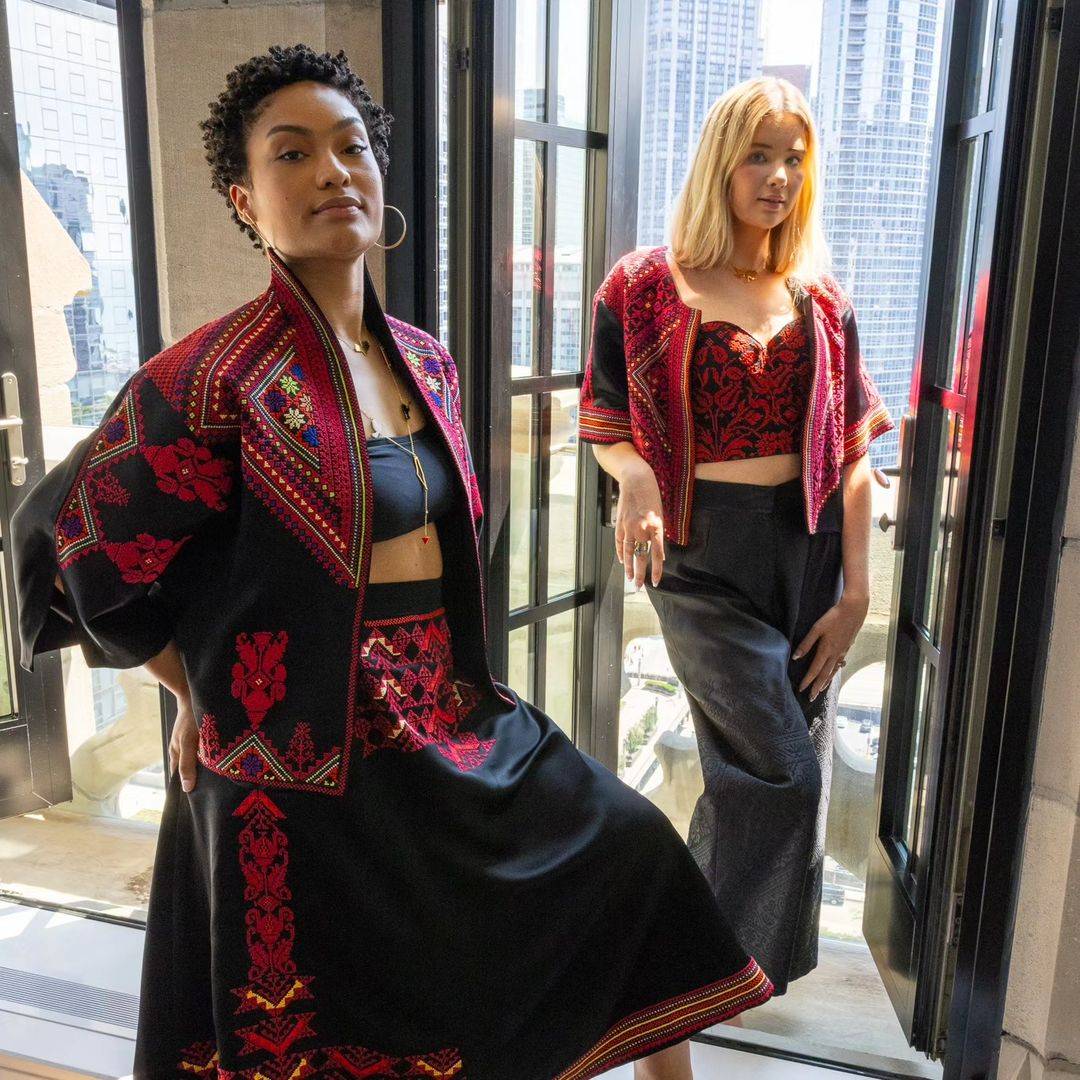 Two models showcasing the Heritage Satin Skirt and matching embroidered jackets, styled with elegant tops, in a modern cityscape setting.