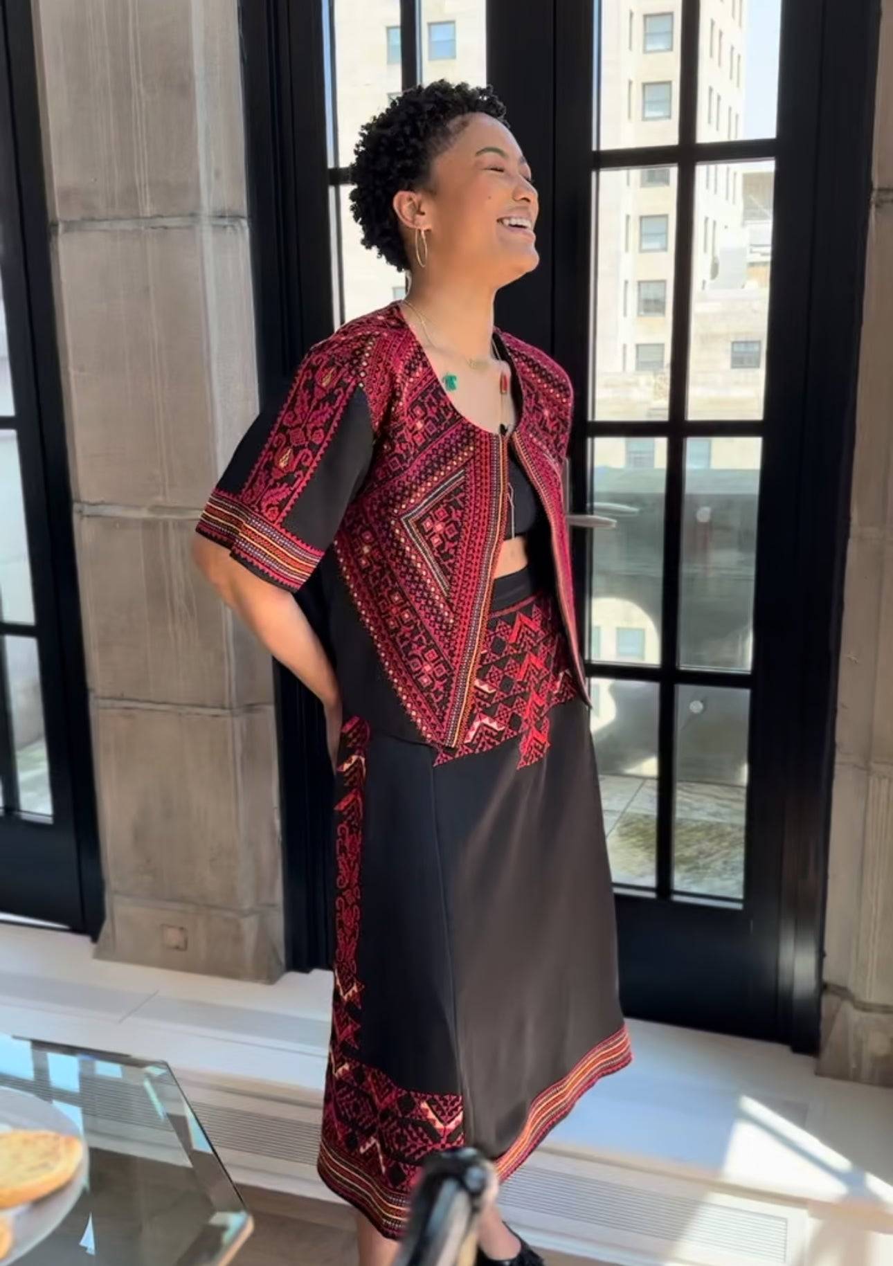 Model wearing the Heritage Satin Skirt with intricate red handcrafted embroidery, paired with a matching embroidered jacket, standing by a window.
